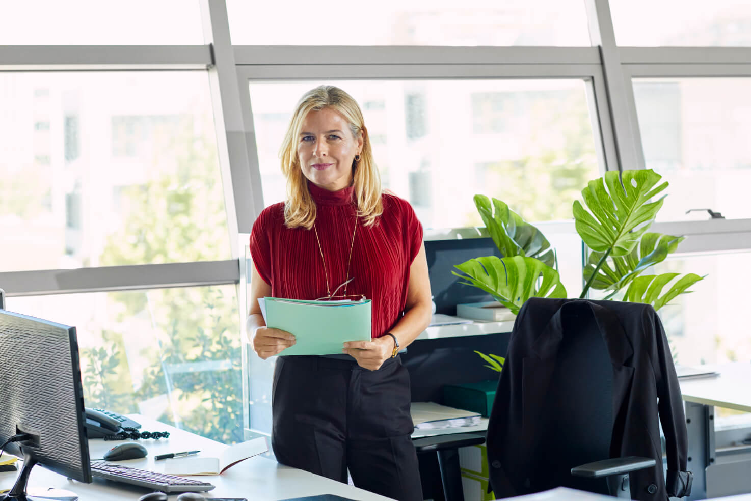 A financial services professional confidently stands next to a desk in an office, holding a folder.